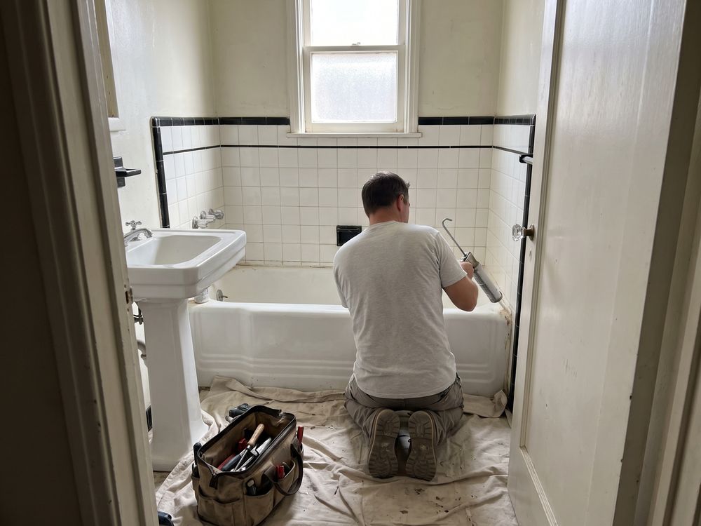 Seen from a bathroom doorway, a handyman kneeling on a drop cloth running a new caulk line along a white tub, his tool bag beside him