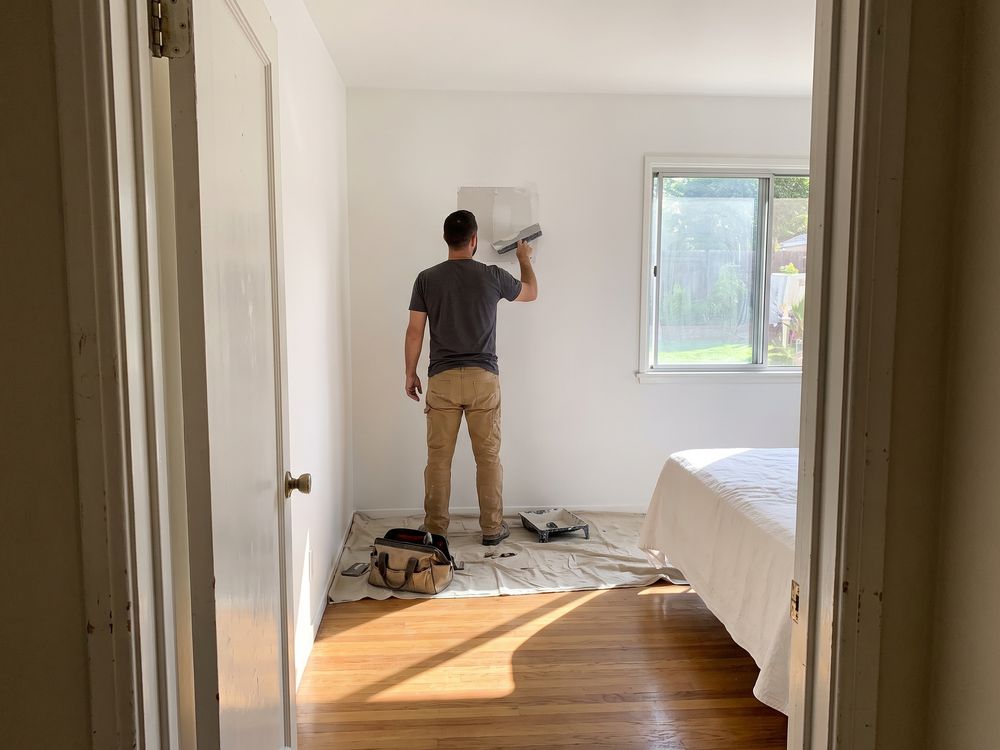 Seen from a bedroom doorway, a handyman with his back turned smoothing a wall patch, his tool bag on a drop cloth at his feet