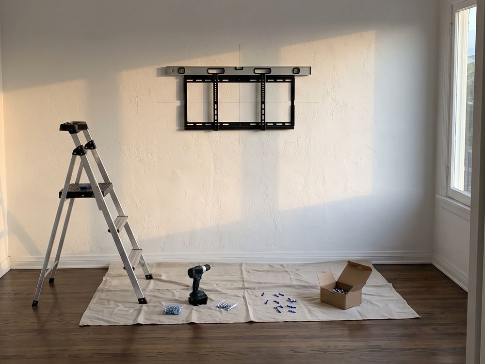 A TV wall plate fixed level on a bare plaster wall, a drop cloth with a drill and anchors on the floor below