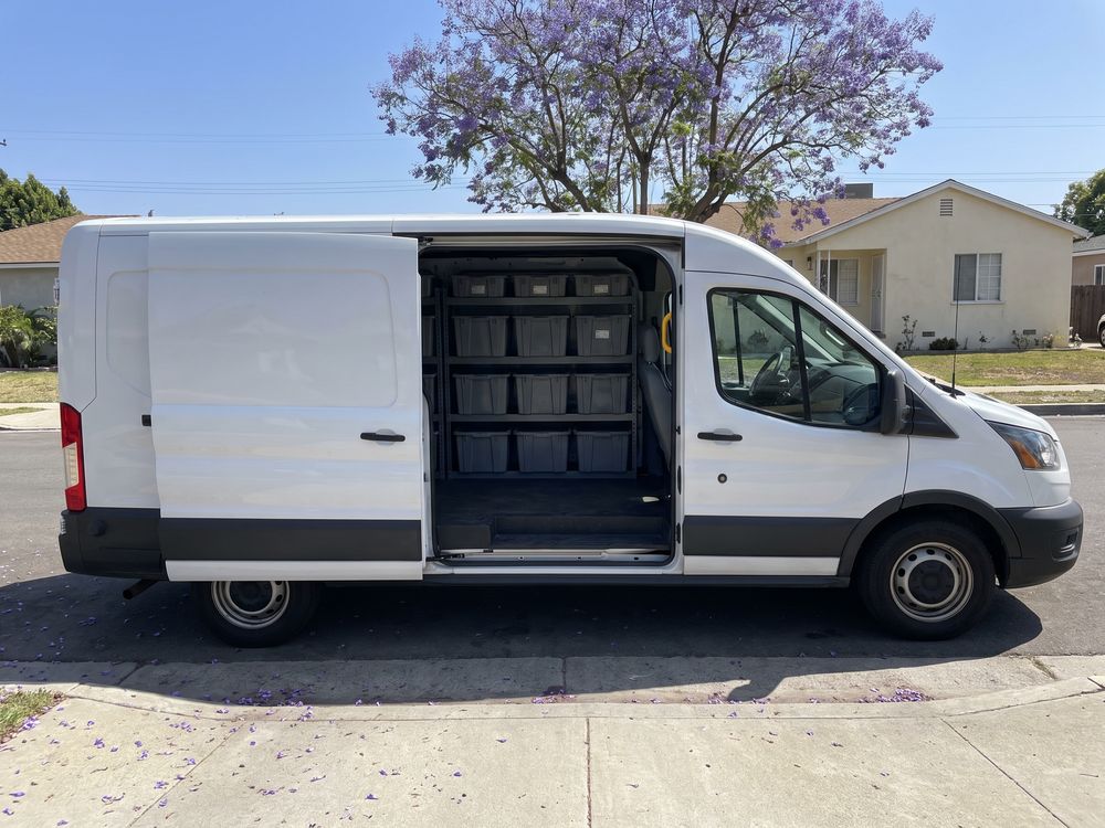 A white work van with its side door open at a Los Angeles curb under a jacaranda tree, parts bins inside
